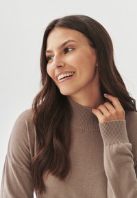 Pull en maille beige avec un col haut à côtes et des manches longues, texture lisse avec des poignets côtelés, design ajusté.