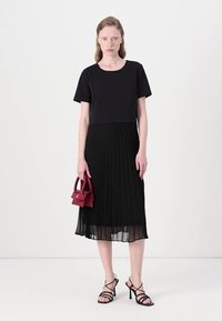 Abito nero a pieghe con parte superiore in tessuto solido, gonna trasparente e maniche corte. Abbinato a tacchi neri con cinturini e una piccola borsa bordeaux.