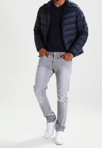 Marinblå pufferjacka, svart V-ringad t-shirt, ljusgrå jeans och vita sneakers. Jackan har dragkedjestängning och quiltat mönster.