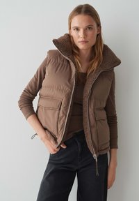 Braune Pufferweste mit einem Kragen aus Kunstfell, ausgestattet mit einem Reißverschluss und seitlichen Taschen. Der Stoff zeigt eine glatte äußere Textur mit gepolstertem Design.