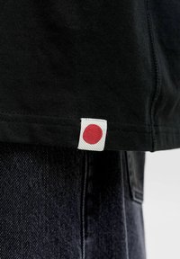 Nahaufnahme des Saums eines schwarzen Hemds mit einem weißen Etikett, das einen roten Kreis zeigt, kombiniert mit dunkler Jeans.