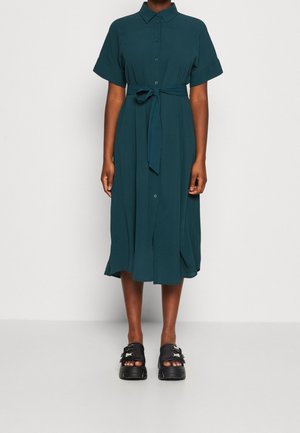 Přiléhavé tyrkysové šaty na knoflíky s límečkem košile, krátkými rukávy a zavázáním v pase. Zvýrazněné rozšířeným lemem. Kombinované s černými sandály na platformě.