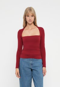 Donna bionda che indossa un top a maniche lunghe bordeaux attillato con scollo quadrato e jeans blu a vita alta, in piedi su uno sfondo bianco.