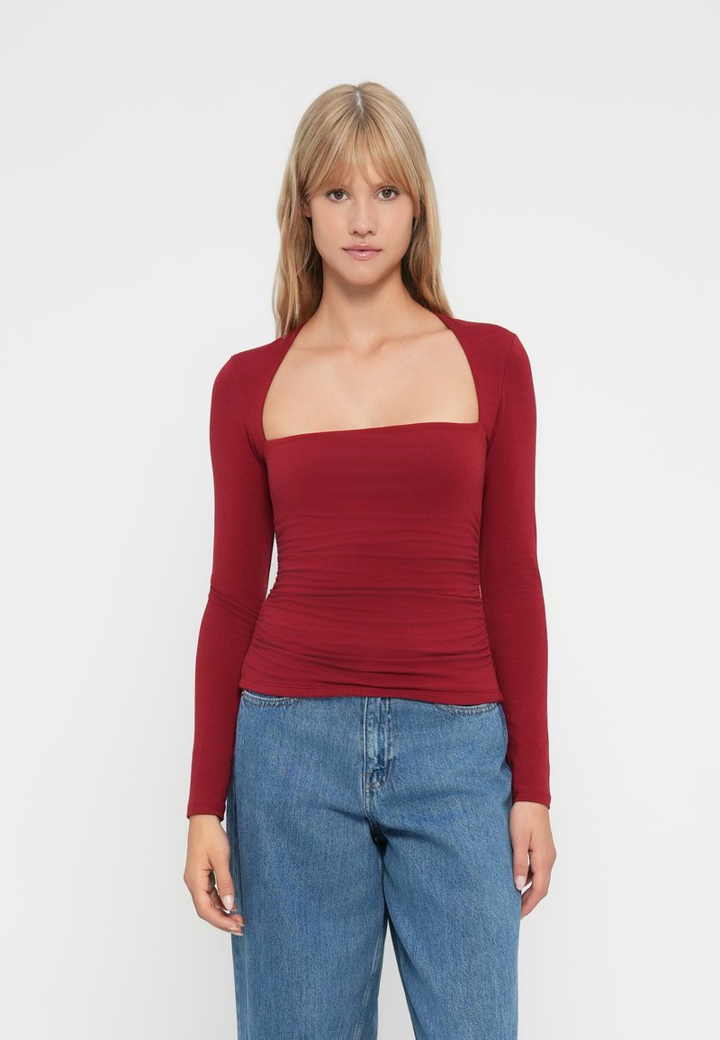 Donna bionda che indossa un top a maniche lunghe bordeaux attillato con scollo quadrato e jeans blu a vita alta, in piedi su uno sfondo bianco.