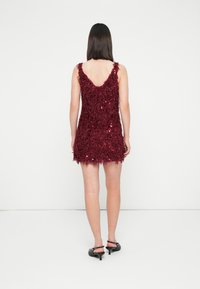 Moteris, aps wearing a belekios, tekstūruotos bordo spalvos mini suknele be rankovių, su juodais mažais kulniukais, stovinti prieš paprastą baltą foną, su nugarą atsukta į priekį.
