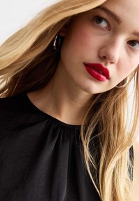 Top nero senza maniche con dettaglio arricciato sul collo, abbinato a grandi orecchini a cerchio in argento. Il modello ha i lunghi capelli biondi e indossa rossetto rosso.