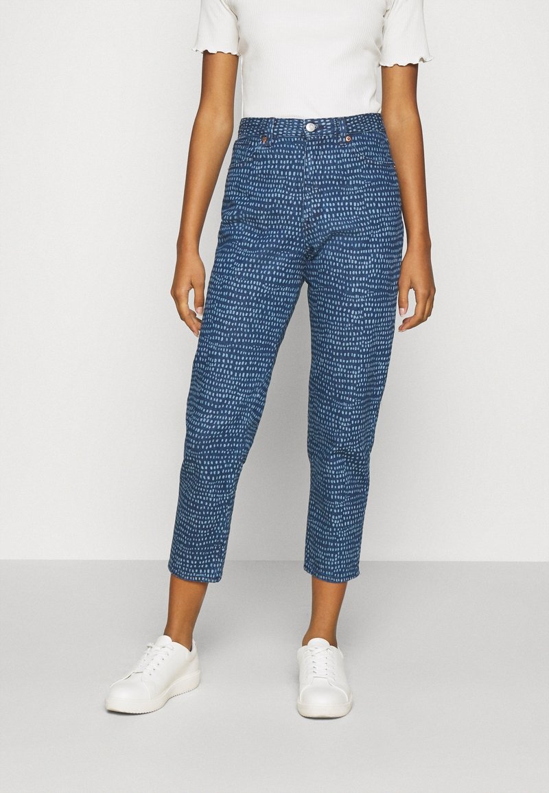 Personne portant un pantalon court bleu taille haute à motif de petits pois blancs, un haut blanc à manches courtes côtelé et des baskets blanches, debout sur un sol gris.