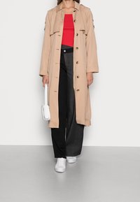 Světle béžový trenčkot s knoflíky, vrstvený přes červený žebrovaný top. V kombinaci s černými širokými kalhotami a bílými teniskami. Drží bílou tašku.