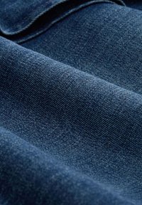 Denim tkanina u dubokoj indigo boji, koja prikazuje glatku teksturu s suptilnim varijacijama u tkanju i preklopljenim rubom.