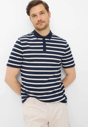 Uomo con capelli castani corti che indossa una maglietta polo a righe orizzontali blu navy e bianca e pantaloni beige chiaro, in piedi contro un muro bianco.