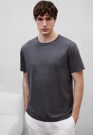 Junger Mann mit kurzen lockigen Haaren, der ein dunkelgraues T-Shirt und weiße Hosen trägt, steht mit den Händen in den Taschen vor einem schlichten Hintergrund.