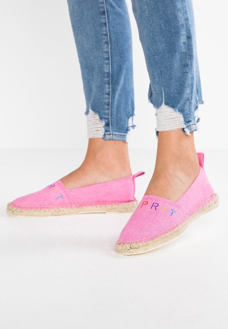 Personne portant des espadrilles roses sans lacets avec des semelles tissées et un logo coloré "ESPRIT", associées à un jean skinny bleu avec ourlet déchiré.