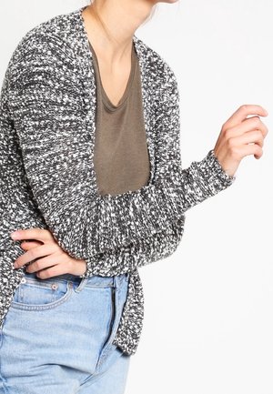 Femme portant un cardigan à motif noir et blanc par-dessus une chemise marron et un jean bleu clair, les bras croisés devant.