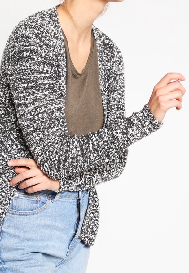 Femme portant un cardigan à motif noir et blanc par-dessus une chemise marron et un jean bleu clair, les bras croisés devant.