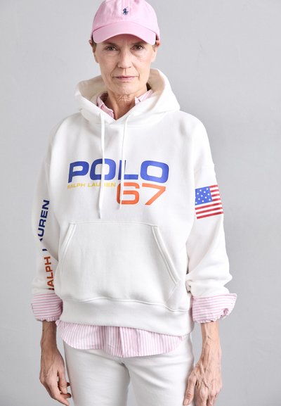 Balts kapucis ar zilu un oranžu "Polo 67" uzrakstu; uz piedurknes ir Amerikas karogs. Valkā virs rozā un balta svītraina krekla. Rozā cepure.