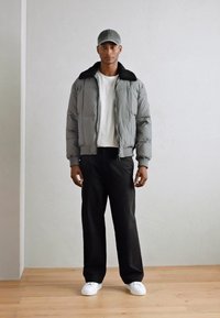 Doudoune grise avec col en polaire noir, t-shirt blanc, pantalon noir et baskets blanches. Le modèle porte une casquette grise.