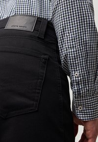 Schwarze Hosen mit strukturiertem Finish, mit einem Lederetikett "pierre cardin", kombiniert mit einem grün-weißen karierten Hemd.