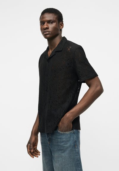 Νεαρός άνδρας στέκεται με το ένα χέρι στην τσέπη του τζιν, φορώντας μια μαύρη δαντελένια μπλούζα με κοντά μανίκια, μπροστά σε ένα απλό φωτεινό φόντο.