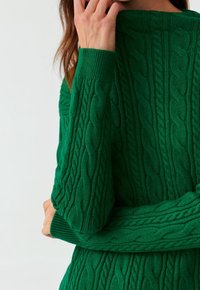 Donna che indossa un maglione verde a treccia con motivi testurizzati, che tiene il mento con una mano, su uno sfondo semplice.