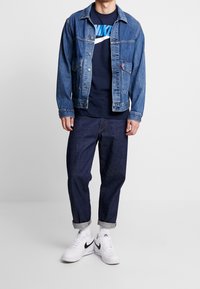 Jeansjacka i ljusblått med två framfickor, marinblå t-shirt med en kraftig blå logotyp, mörkblå uppvikta byxor och vita sneakers.