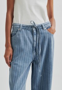 Világoskék farmernadrág fehér függőleges csíkokkal, állítható derékkal, oldalsó zsebekkel és laza szabással.