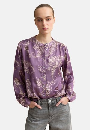 Mujer joven con el cabello peinado hacia atrás, vistiendo una blusa morada con botones y diseño de cachemira y pantalones vaqueros gris claro, con las manos en los bolsillos.