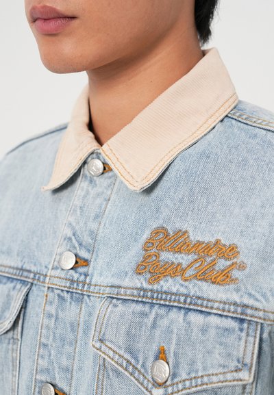 Džinsa jaka ar gaiši zilu krāsu, beidža krāsas kordūrā apmalēm, sudraba pogām un zelta izšūtu "Billionaire Boys Club" logo.