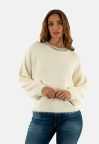 Maglione crema con una finitura texturizzata e fuzzy, e un collo rotondo decorato con piccole perline trasparenti. Vestibilità ampia e maniche lunghe, abbinato a jeans blu.