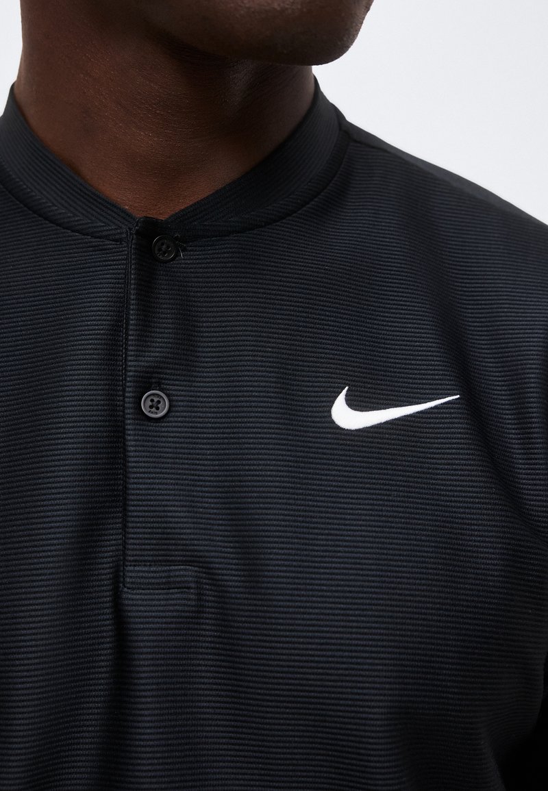 Gros plan d'une personne portant un t-shirt Nike noir côtelé avec deux boutons noirs et le logo Nike Swoosh blanc sur la poitrine.