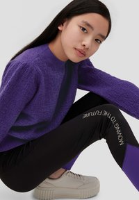 Pull en maille violet, leggings noirs avec l'imprimé "PASSER À L'AVENIR", et baskets beige clair avec des semelles épaisses.