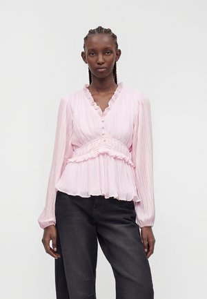 Femme aux cheveux tressés portant un chemisier plissé rose clair avec bordure volantée et un pantalon noir sur un fond blanc uni.
