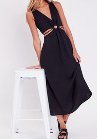 Robe maxi noire sans manches avec un profond décolleté en V et un détail noué à la taille, présentant des découpes et un accent circulaire. Texture lisse.