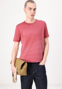 Rød T-skjorte med korte ermer, teksturert stoff og en subtil Diesel-logo-print. Kombinert med mørke jeans og en lys messenger bag.