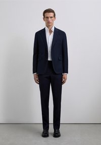 Chaqueta de traje azul marino, camisa blanca y pantalones oscuros. Tela lisa con corte entallado, solapas con muesca y detalles de costura sutiles. Zapatos negros.