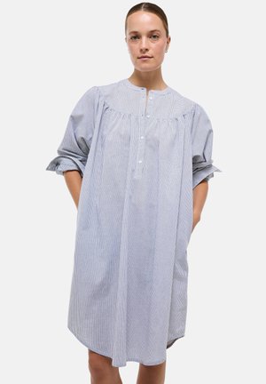 Femme aux cheveux attachés portant une robe ample à manches longues boutonnée, à carreaux gris, avec un empiècement froncé et les mains dans les poches.