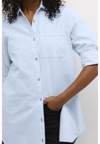 Chemise bleu clair à boutons, en tissu texturé. Comprend une poche poitrine, des manches retroussées et un ourlet arrondi.