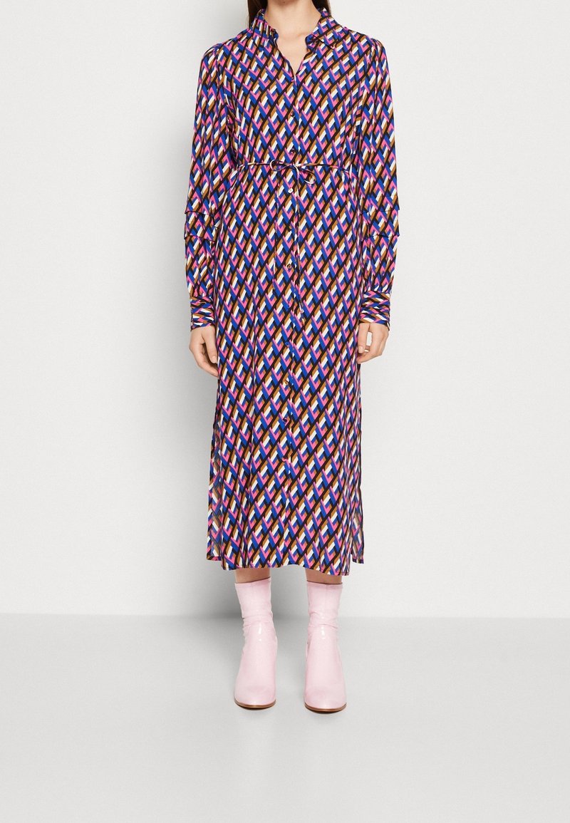 Langärmliges, knopfbares Kleid mit einem Rautenmuster in Lila, Rosa und Blau. Verfügt über eine gebundene Taille und wird mit rosa Absatzstiefeln kombiniert.