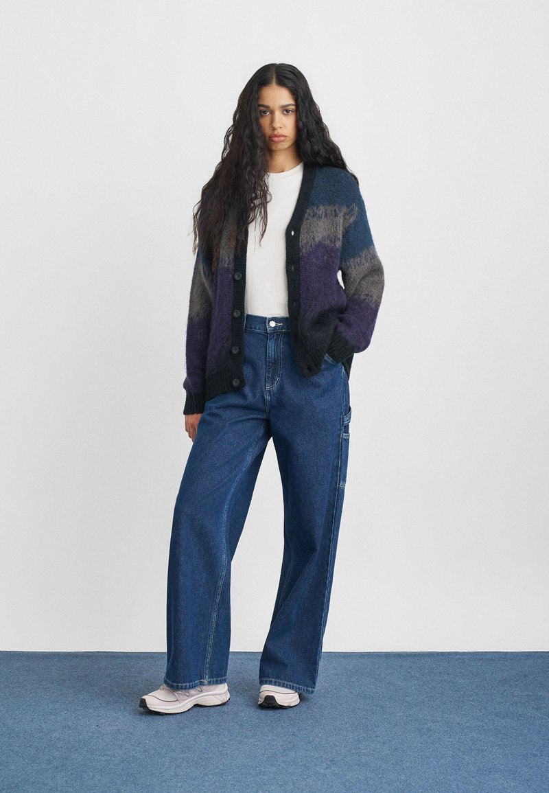 Woman with long curly hair wearing a multicolor buttoned cardigan, white shirt, wide blue jeans, and light-colored sneakers standing on blue carpet.