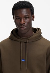 Barna kapucnis pulóver zsinórokkal és egy kis kék téglalap alakú logóval, amelyen a "HUGO" felirat olvasható a bal mellkason. Sima tapintású és laza szabású.