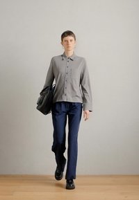 Γκρίζο πουκάμισο με κουμπιά και μακριά μανίκια, ελαφρύ ύφασμα και μικρή τσέπη στο στήθος; συνδυασμένο με ναυτικά παντελόνια και μαύρα παπούτσια; κρατώντας μαύρη τσάντα.