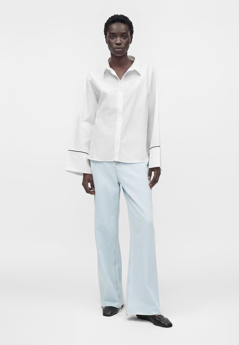 Femme portant une chemise blanche oversize avec de larges poignets et un jean large bleu clair, debout devant un fond clair uni.