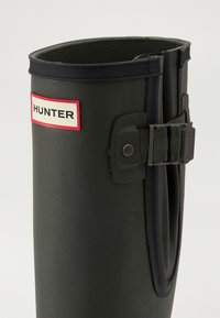Botín de goma verde con acabado texturizado, presenta un borde negro, correa ajustable y un logo visible de "HUNTER" en rojo y blanco.