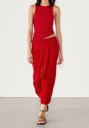 Rød strikket to-delt sæt bestående af en ærmeløs top med asymmetrisk snit og en draperet nederdel med knuddet detalje. Sorte strapsandaler.