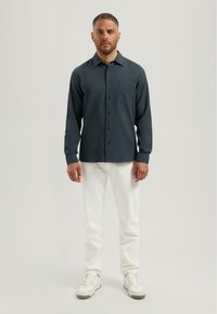 Donkergroene langemouwen shirt van een textuurstof, met een voorzak en een button-down kraag, gecombineerd met witte broek en lichte sneakers.