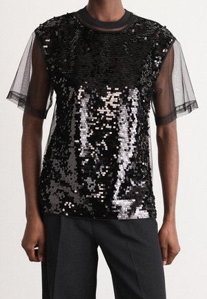 Czarna cekinowa bluzka z przezroczystymi krótkimi rękawami, noszona z czarnymi spodniami, pokazana od barków do bioder na stojącej osobie.