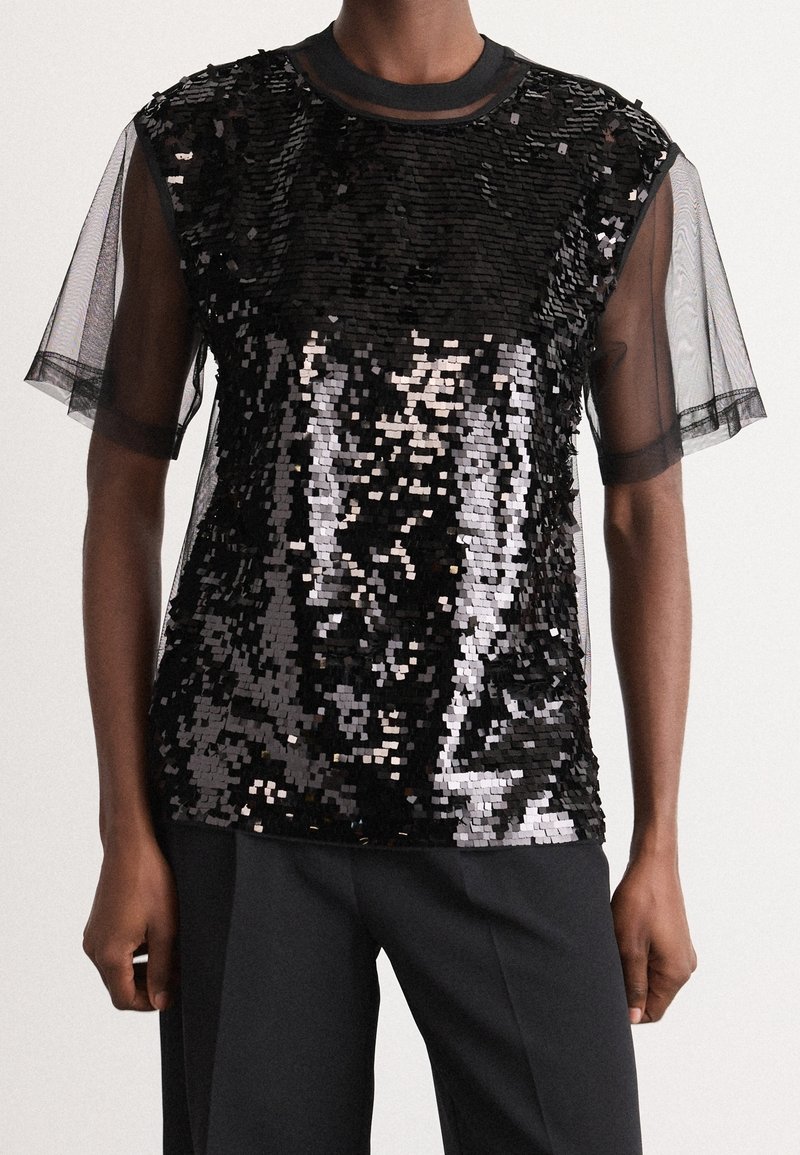 Haut noir à sequins avec manches courtes transparentes porté avec un pantalon noir, vu des épaules aux hanches sur une personne debout.