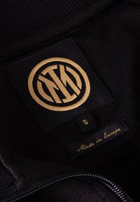 Zwarte stoffen label met een gouden, cirkelvormig logo, maat "S" en de tekst "Made in Europe"; heeft een gestructureerde rand en een glad oppervlak.