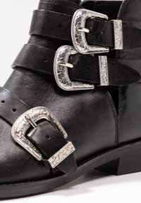 Černá kožená kotníková bota s třemi širokými pásky a zdobenými stříbrnými sponami, s mírným podpatkem. Hladká textura a bezpečný design.