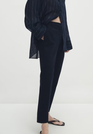 Personne portant une chemise noire à fines rayures et manches longues, un pantalon noir droit et des tongs noires, debout de profil avec la main dans la poche.