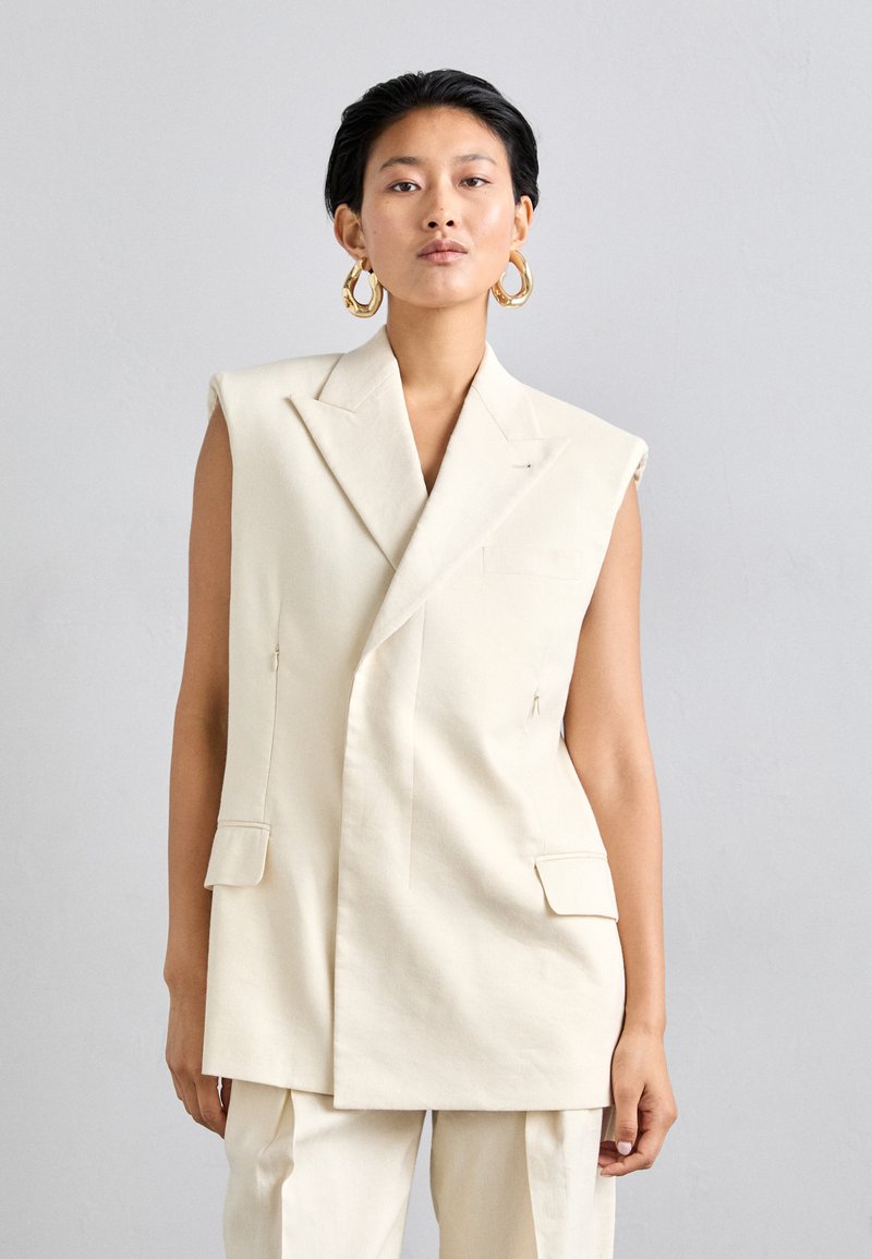 Blazer sans manches en ivoire, fabriqué dans un tissu structuré, avec un design croisé, deux poches avant et des détails de couture subtils.